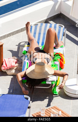 Die Insel Santorini. Gegerbten jungen kaukasischen schlanke Frau Verlegung auf Sonnenliege mit schwarzen Bikini an und Strohhut über über Gesicht gezogen. Ansicht von oben. Stockfoto