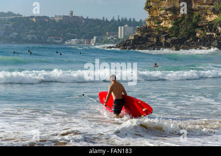 Ein Mann mit einem roten Stand-up-Paddleboard (SUP) steigt an einem sonnigen Sydney-Morgen in Australien in die Brandung am Freshwater Beach ein Stockfoto