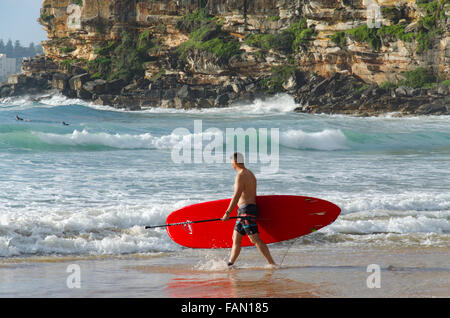 Ein Mann mit einem roten Stand-up-Paddleboard (SUP) steigt an einem sonnigen Sydney-Morgen in Australien in die Brandung am Freshwater Beach ein Stockfoto