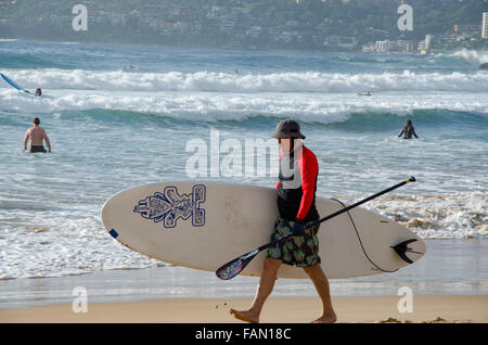 Ein älterer Mann, der an einem sonnigen Morgen in Sydney in Australien ein Stand-up-Paddleboard (SUP) am Freshwater Beach trägt Stockfoto