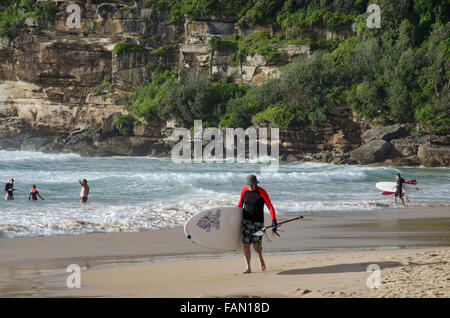 Ein älterer Mann, der an einem sonnigen Morgen in Sydney in Australien ein Stand-up-Paddleboard (SUP) am Freshwater Beach trägt Stockfoto