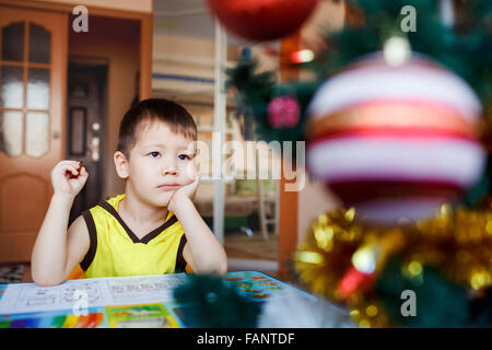 Verträumter kleine Junge sitzt auf einem Hintergrund Weihnachtsbaum schreibt seiner Wunschliste. Stockfoto