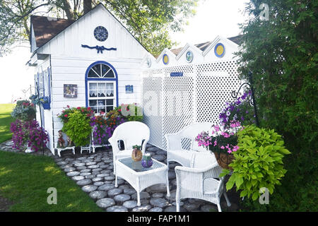 Ein schönes kleines Gartenhäuschen mit schönen Stühlen und vielen Blumen im Garten, neu in weiß lackiert. Stockfoto