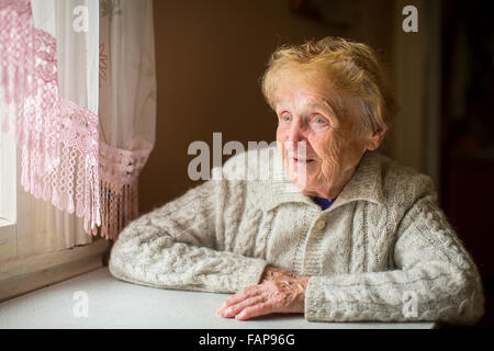 Eine ältere Frau sitzt an einem Tisch in der Küche neben dem Fenster. Stockfoto