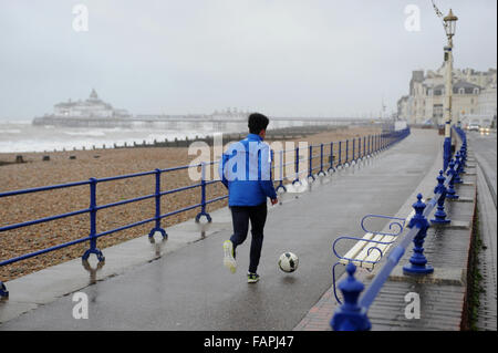 Ein junger Mann tritt einen Fußball entlang der Promenade an einem nassen und windigen Tag in Eastbourne, East Sussex, England. Stockfoto