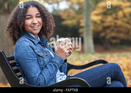 Schöne glückliche Mischlinge afroamerikanische Mädchen Teenager weibliche junge Frau lächelt trinken Kaffee draußen sitzen auf Bank Stockfoto