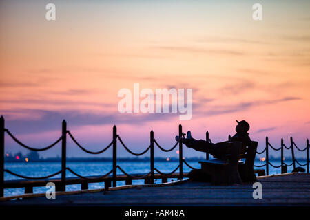 Ein Mann sitzt Liegeposition, Silhouette gegen den Sonnenuntergang entlang der Uferpromenade. Stockfoto