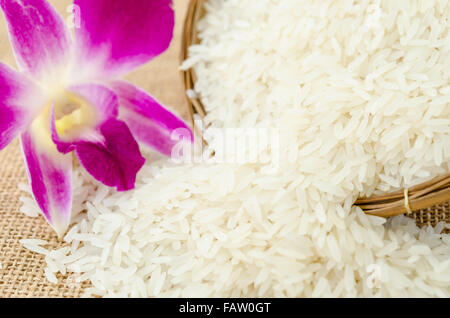 Roher Reis mit violetten Orchidee Blume im Weben Korb auf Sack Hintergrund. Stockfoto