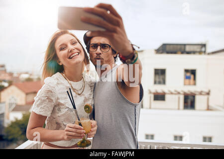 Zwei junge Freunde, die auf dem Dach eine Selfie übernehmen. Mann hält Smartphone und Selbstportrait mit Frau hält einen cocktail dur Stockfoto