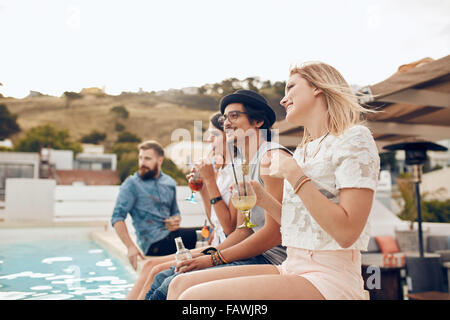 Glückliche junge Freunde sitzen am Pool mit cocktail. Junge Menschen entspannen am Pool mit den Füßen im Wasser dur Stockfoto