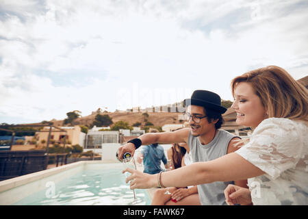 Junger Mann gießt Champagner im Glas von seiner Freundin während der Party am Pool. Junge Menschen, die Spaß am Dach p Stockfoto
