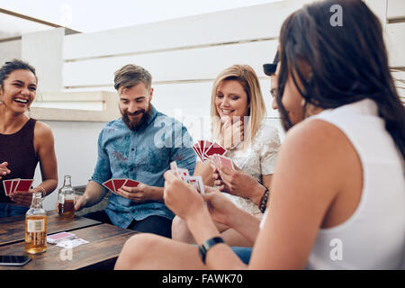 Gruppe von Freunden sitzen an einem Holztisch und Spielkarten. Junge Menschen spielen eine Partie Karten während einer Party. Stockfoto