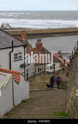 Zwei Menschen, die zu Fuß einen Hund auf eine Straße hinunter in Richtung Hafen von Whitby, England Stockfoto