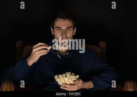 junger Mann sitzt im dunklen Zimmer an der Vorderseite tv Film und Essen popcorn Stockfoto