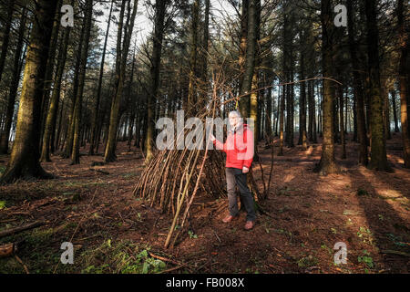 Leuchtfeuer fiel, Lancashire, England. Mann in rot Outdoorjacke neben dem teilweise Stand gebaut Tierheim gemacht aus Stöcken und Zweigen. Stockfoto