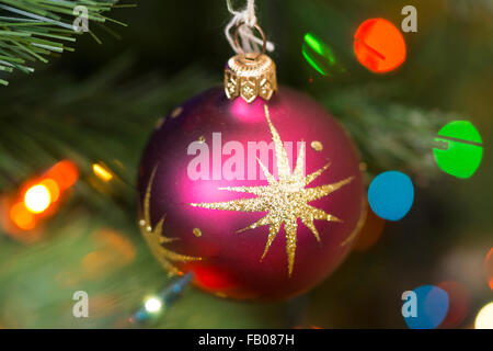 Eine rosa Weihnachtskugel mit goldgelben Glitzersternen, die an einem Weihnachtsbaum hängen und von Lichterketten beleuchtet werden Stockfoto