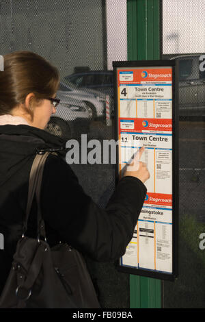 Eine junge Frau in ihren Zwanzigern mit Mantel und Handtasche, die einen Busfahrplan konsultiert und auf 'Town Center' an einer Bushaltestelle, Basingstoke, Großbritannien, zeigt Stockfoto