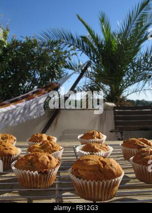Hausgemachte Kürbis-Walnuss-Muffins am Kühlregal platziert auf einem Holztisch in der Sonne vor einer Palme und Hängematte Stockfoto