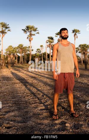 Voller Länge Vorderansicht der junge Mann stand vor Palmen werfen Schatten wegsehen, Taiba, Ceara, Brasilien Stockfoto