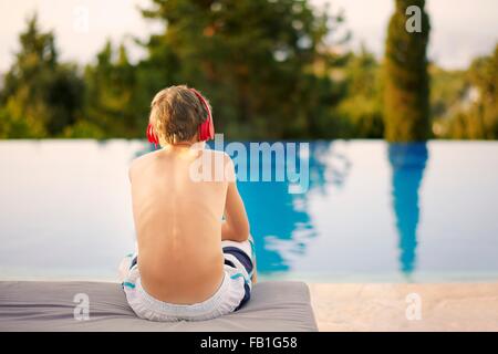 Rückansicht des Teenager tragen Kopfhörer sitzt am Infinity-pool Stockfoto