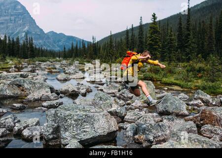 Seitenansicht der Mitte erwachsener Mann mit Rucksack springen über felsigen Flussbett, Moraine Lake, Banff Nationalpark, Alberta Kanada Stockfoto