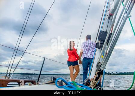 Rückansicht des Paares am Bug des Bootes, die Hand in Hand, Blick über die Schulter in die Kamera Lächeln Stockfoto