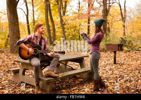 Junge Frau fotografieren Gitarrenspiel Freund im herbstlichen Wald Stockfoto
