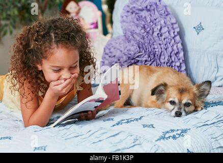 Mädchen und Hund auf Bett Buch zu lesen, hand auf Mund lachen Stockfoto