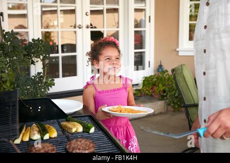 Mädchen auf der Terrasse halten Platte wird Speisen Grill von Vater Stockfoto