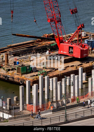 Ein Kran auf einem Lastkahn hebt Pfähle für den Bau der Küstenlinie in den Hudson River neben der Lower West Side von Manhattan, New York City. Beachten Sie nummerierte Pfähle bereits im Ort und Fußgänger am Riverwalk. Stockfoto