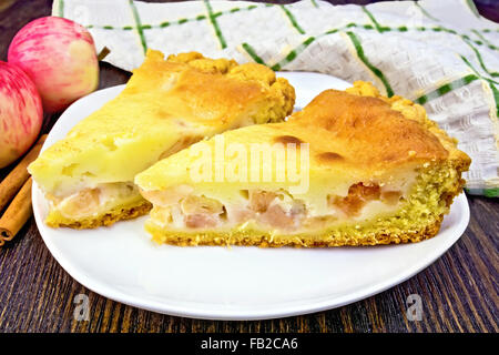 Zwei Stück Apfelkuchen mit Sahne-Sauce in weißer Teller, Zimt, Äpfel und eine Serviette auf einem dunklen Holzbrett Stockfoto