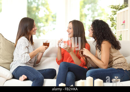 Drei glückliche Freunde reden und trinken Kaffee und Tee auf der Couch zu Hause sitzen Stockfoto