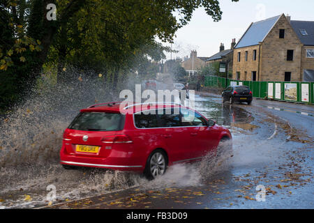 Nach starken Regenfällen fährt Auto durch große Pfütze auf überfluteten Straße, Besprühen mit Wasser wie es Kurve ins Dorf Runden. Burley in Wharfedale, GB, UK. Stockfoto