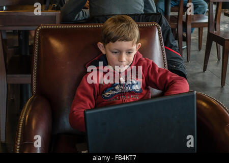 Technisch versierte junge 10-12 Jahre alt, sitzt in einem Café mit einem großen Laptop auf seinem Schoß, wie konzentriert er sich auf sie. Stockfoto