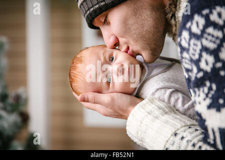 Vater Holding und küssen Baby Sohn unter während des Tragens winter Hut, mit Weihnachten Baum im Hintergrund Stockfoto