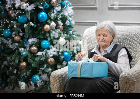 Ältere Frau öffnet eine Paket mit einem Geschenk beim sitzen neben einem Weihnachtsbaum Stockfoto