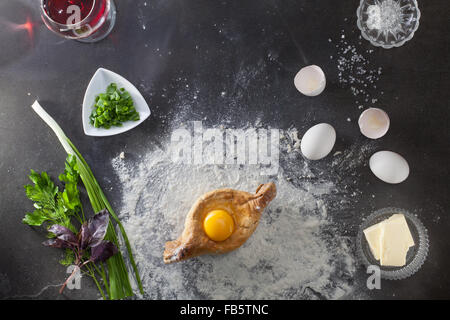 Khachapuri auf schwarzen Tisch mit Mehl und Zutaten. Ansicht von oben Stockfoto