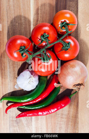 Fünf Strauchtomaten, Knoblauchzwiebel, eine Zwiebel und vier Chilischoten auf ein Schneidebrett von oben Stockfoto