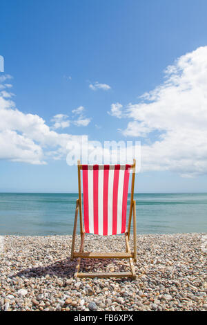 Eine einzelne rote und weiß gestreiften Liegestuhl mit Blick auf den Ozean auf einer einsamen Kieselstrand. Stockfoto