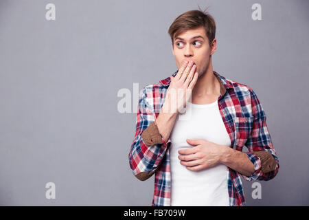 Erstaunt erstaunt jungen Mann im karierten Hemd bedeckt Mund mit der Hand und Gefühl Angst über grauen Hintergrund Stockfoto