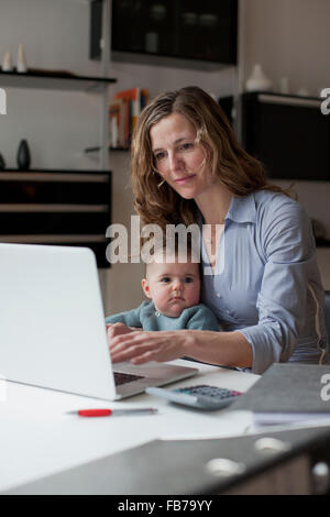 Frau auf Laptop sitzend mit Baby Girl zu Hause arbeiten Stockfoto