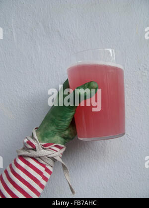 Nahaufnahme von Grün bemalte Hand mit Glas voller rotes Getränk gegen weiße Wand Stockfoto