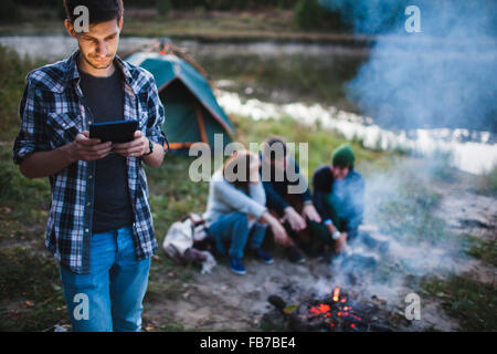 Junger Mann mit digital-Tablette während Freunde sitzen Lagerfeuer im Wald Stockfoto