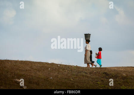 Eine Frau, die einen Plastikeimer auf dem Kopf trägt, während sie mit Kindern während der Trockenzeit auf Sumba Island, Indonesien, auf trockener Landschaft läuft. Stockfoto