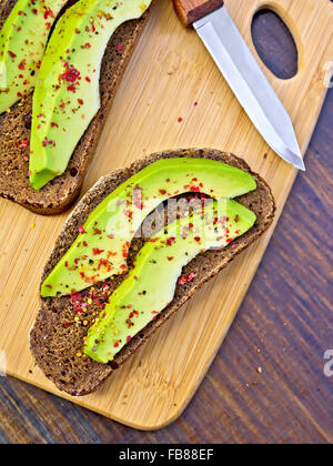 Zwei Scheiben Graubrot mit Scheiben von Avocado und Pfeffer, Messer gegen die dunkle Holzplatte Stockfoto