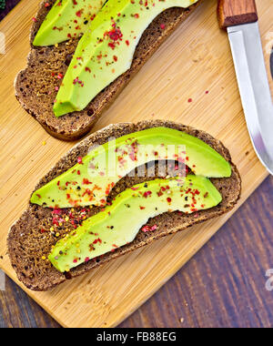 Zwei Scheiben Graubrot mit Scheiben von Avocado und Pfeffer, Messer auf dunklen Holzbrettern Hintergrund Stockfoto