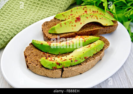 Zwei Scheiben Graubrot mit Scheiben von Avocado und Pfeffer auf einem weißen Teller, grüne Serviette, Petersilie auf dem Hintergrund Licht aus Holz Stockfoto