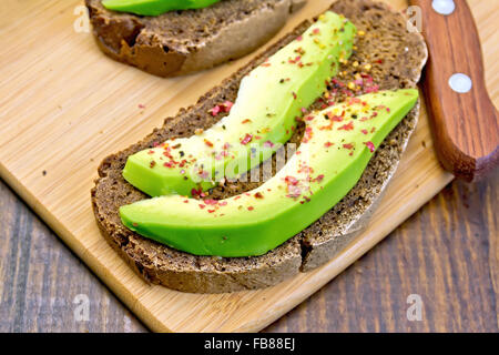 Zwei Scheiben Graubrot mit Scheiben von Avocado und Pfeffer, Messer gegen die dunkle Holzplatte Stockfoto