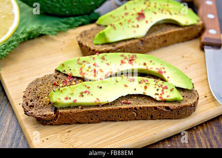 Zwei Scheiben Graubrot mit Scheiben von Avocado und Pfeffer, Zitrone, Serviette, Messer gegen die dunkle Holzplatte Stockfoto