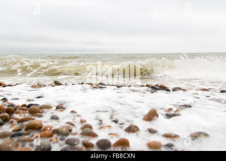 Schäumende Wellen wäscht und stürzt über Kieselsteine am britischen Küste Kiesstrand an einem grauen, bewölkten Tag Stockfoto